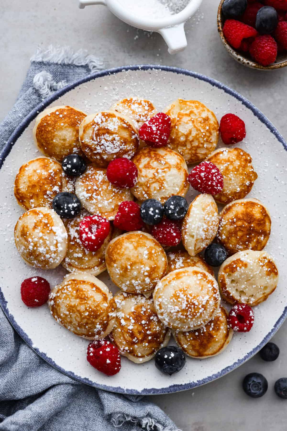 Poffertjes are small, fluffy Dutch pancakes that are so delicious for breakfast! Serve them with a dusting of powdered sugar and fresh berries for a meal that everyone will love. Poffertjes served with berries and a dusting of powdered sugar on a blue and white plate.