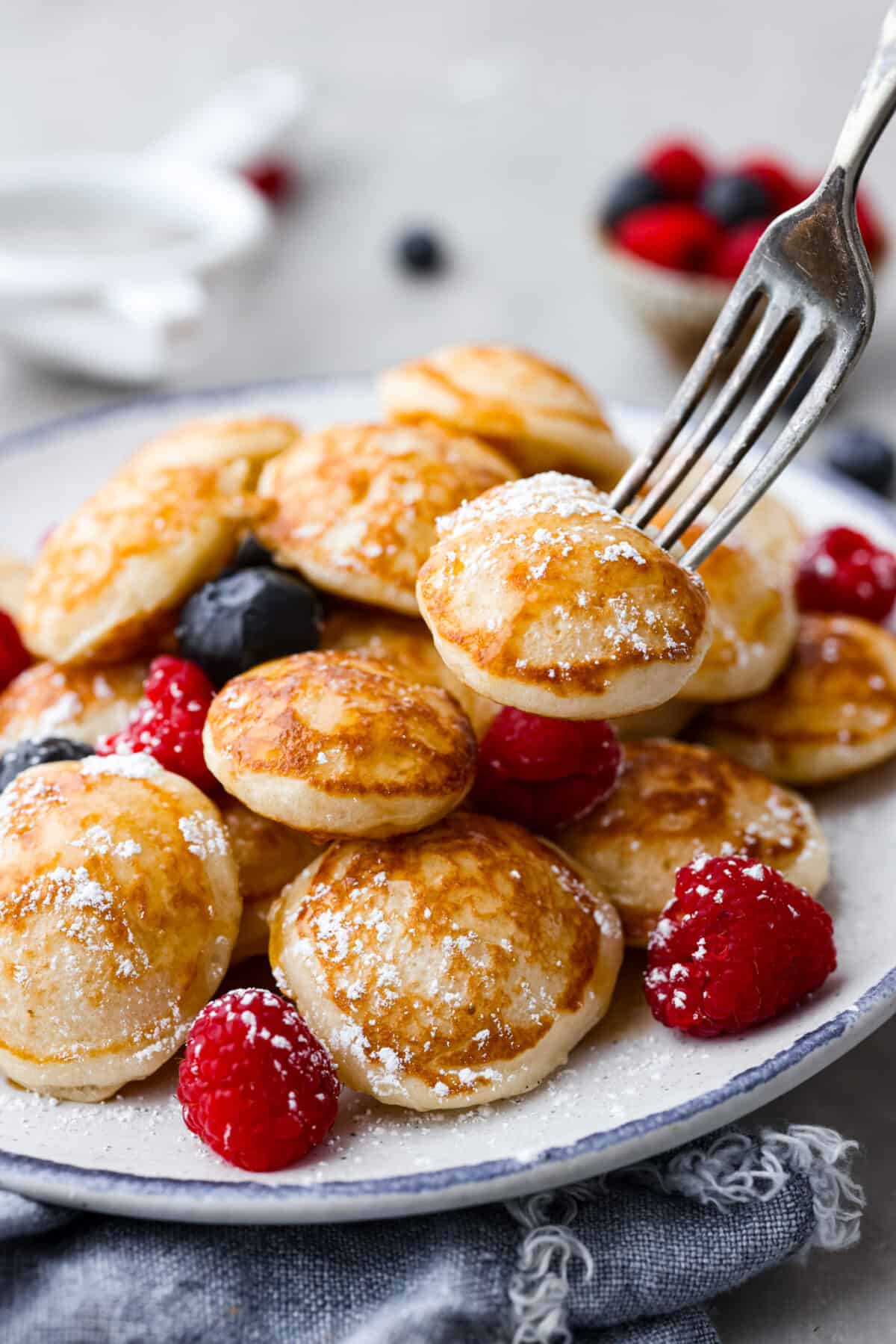 Poffertjes are small, fluffy Dutch pancakes that are so delicious for breakfast! Serve them with a dusting of powdered sugar and fresh berries for a meal that everyone will love. Closeup of small Dutch pancakes served with berries and powdered sugar.