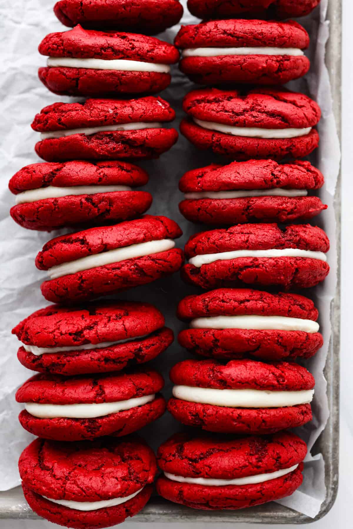 These vibrant red velvet Oreos are filled with a sweet cream cheese frosting and are irresistibly cute! Super easy to make from a red velvet cake mix, but SO delicious that no one will ever know. Overhead view of red velvet Oreos on a baking sheet pan lined with parchment paper.