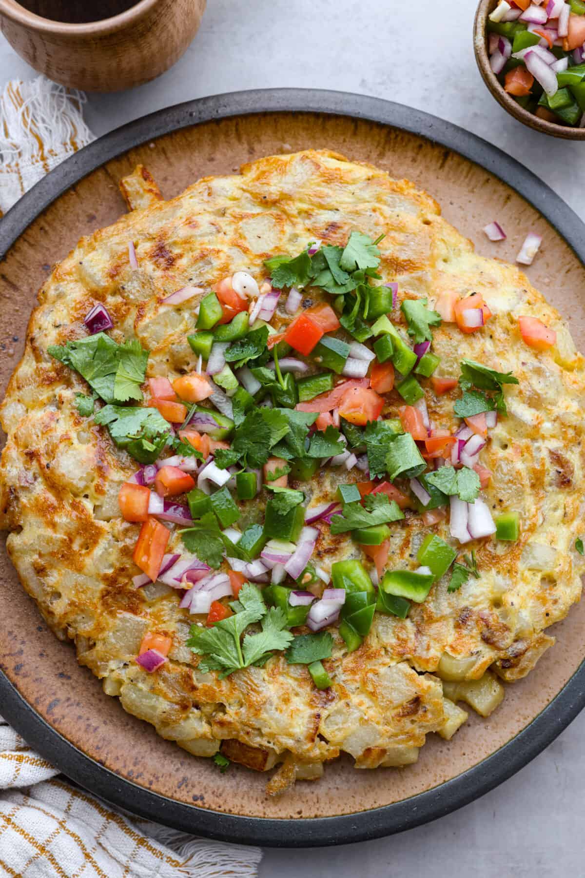 Make this delicious traditional Spanish tortilla at home! Tender potatoes, eggs, and onions cooked together in a skillet and topped with fresh salsa. Bring the flavors of Spain to your kitchen! Top view of Spanish tortilla on a large plate topped with salsa and cilantro.