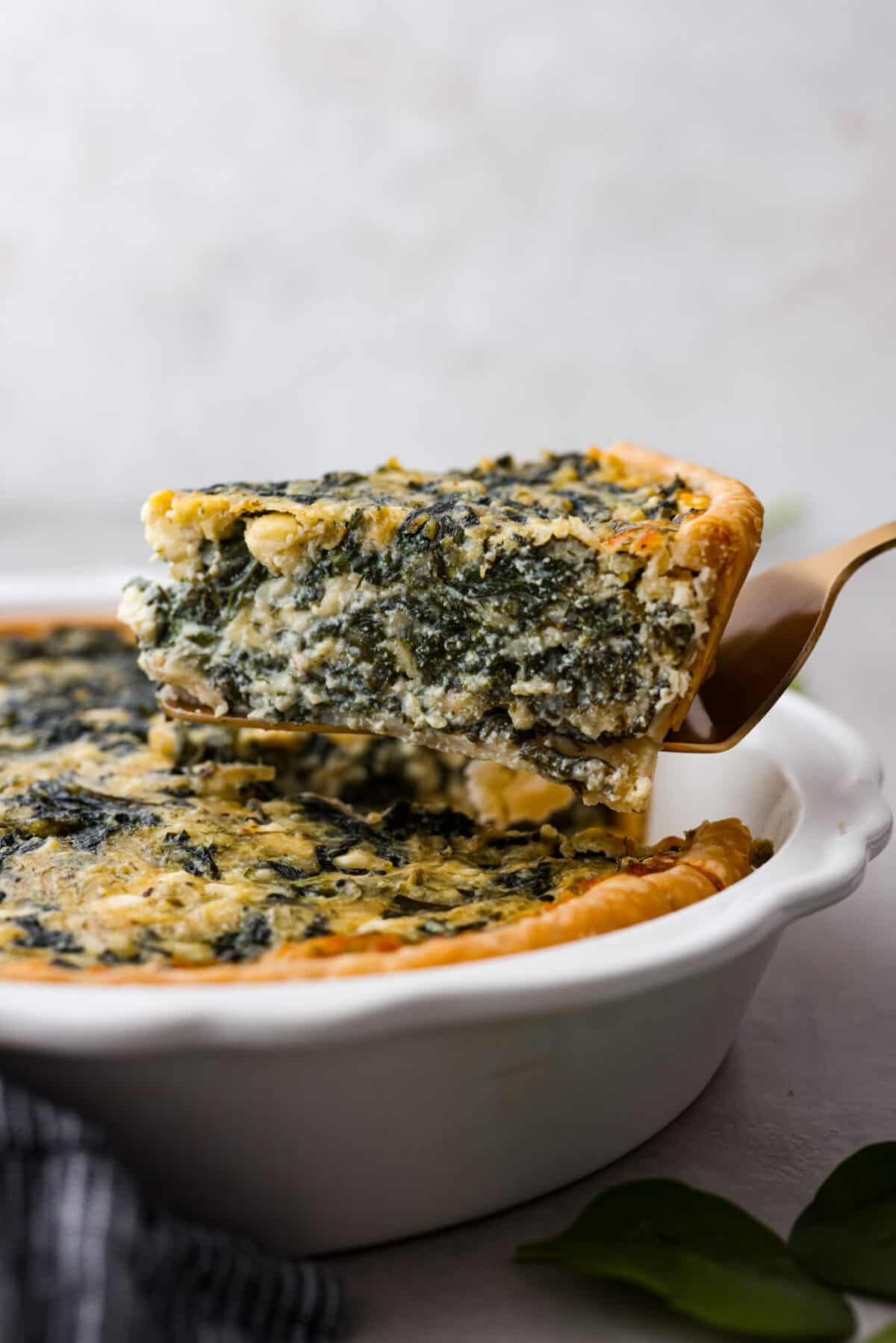 This savory spinach pie is packed with spinach and baked in a flaky crust. It's loaded with three kinds of cheeses, onion, garlic, eggs, and seasoned with tons of flavor! Enjoy it warm or cold, and any time of day. Close side view of a gold serving spatula holding up a slice of pie.