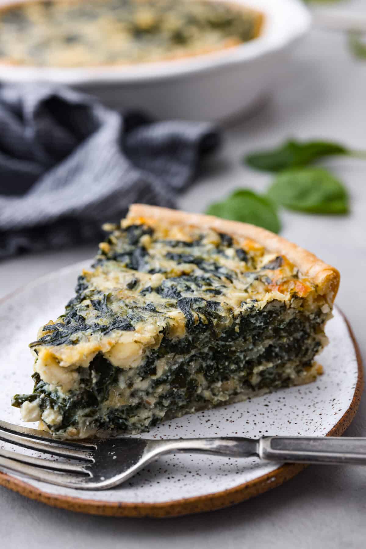 This savory spinach pie is packed with spinach and baked in a flaky crust. It's loaded with three kinds of cheeses, onion, garlic, eggs, and seasoned with tons of flavor! Enjoy it warm or cold, and any time of day. Close view of a slice of spinach pie on a small white plate with a fork.