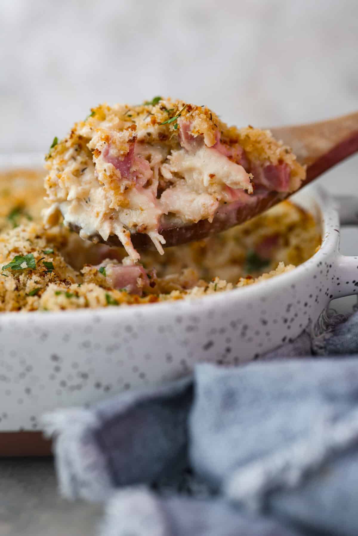 This Chicken Cordon Bleu Casserole is simple and delicious. It combines tender chicken and smokey, flavorful ham in a melty, creamy cheese sauce topped with crispy breadcrumbs. Your whole family will love it! A scoop of chicken cordon bleu casserole on a wooden serving spoon.
