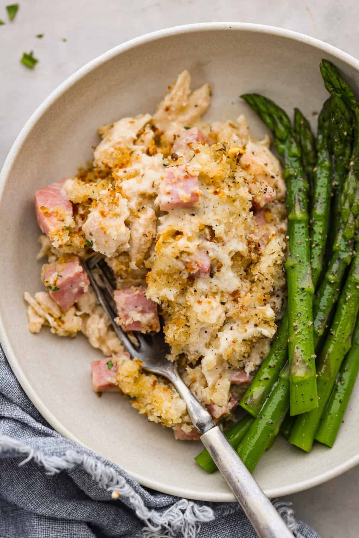 This Chicken Cordon Bleu Casserole is simple and delicious. It combines tender chicken and smokey, flavorful ham in a melty, creamy cheese sauce topped with crispy breadcrumbs. Your whole family will love it! A scoop of the casserole served with asparagus in a cream-colored dish.