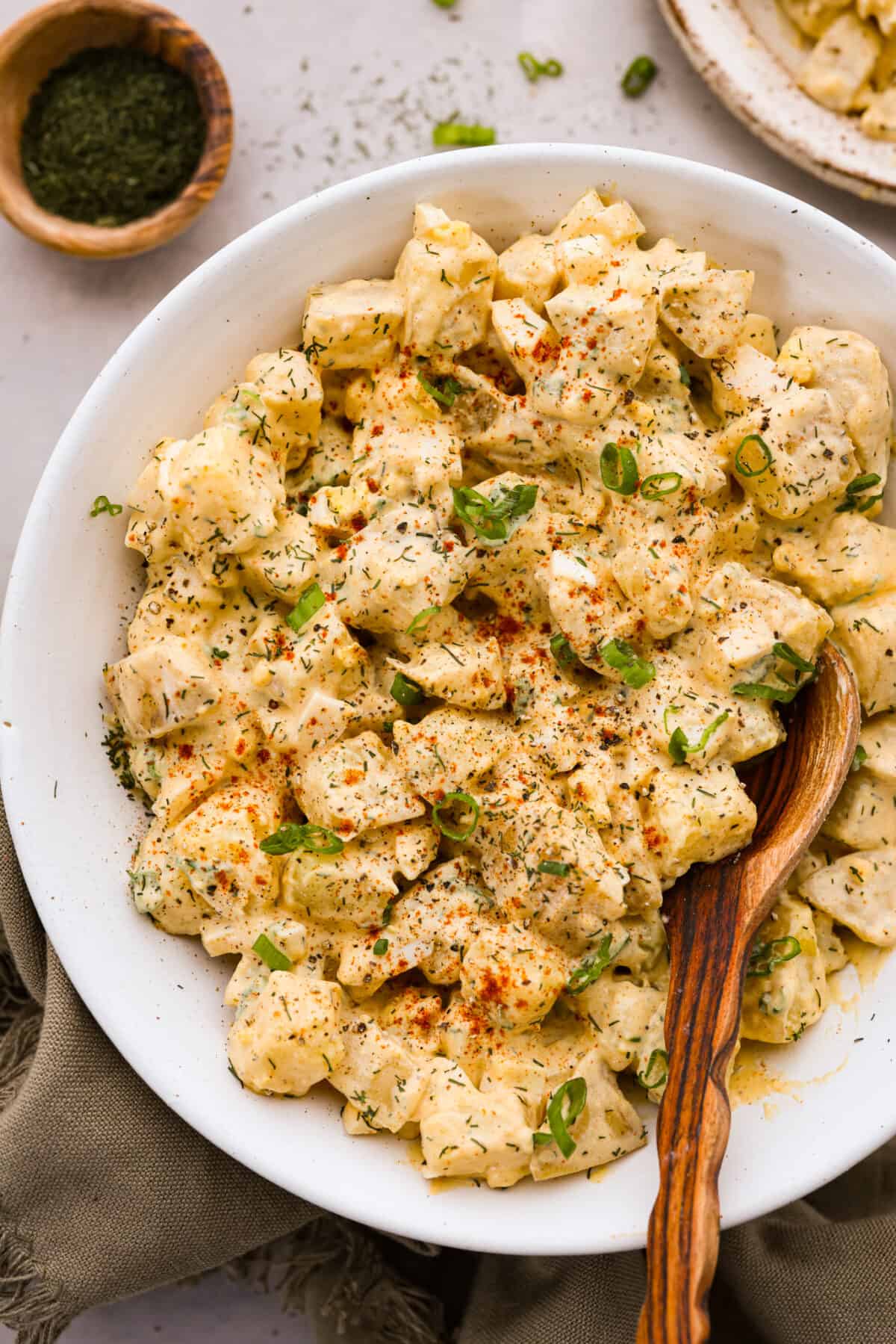 This classic potato salad recipe is cool, creamy, and the perfect side dish for any cookout! Easy to make ahead and so flavorful! Potato salad served in a white bowl and garnished with green onions, paprika, and herbs.