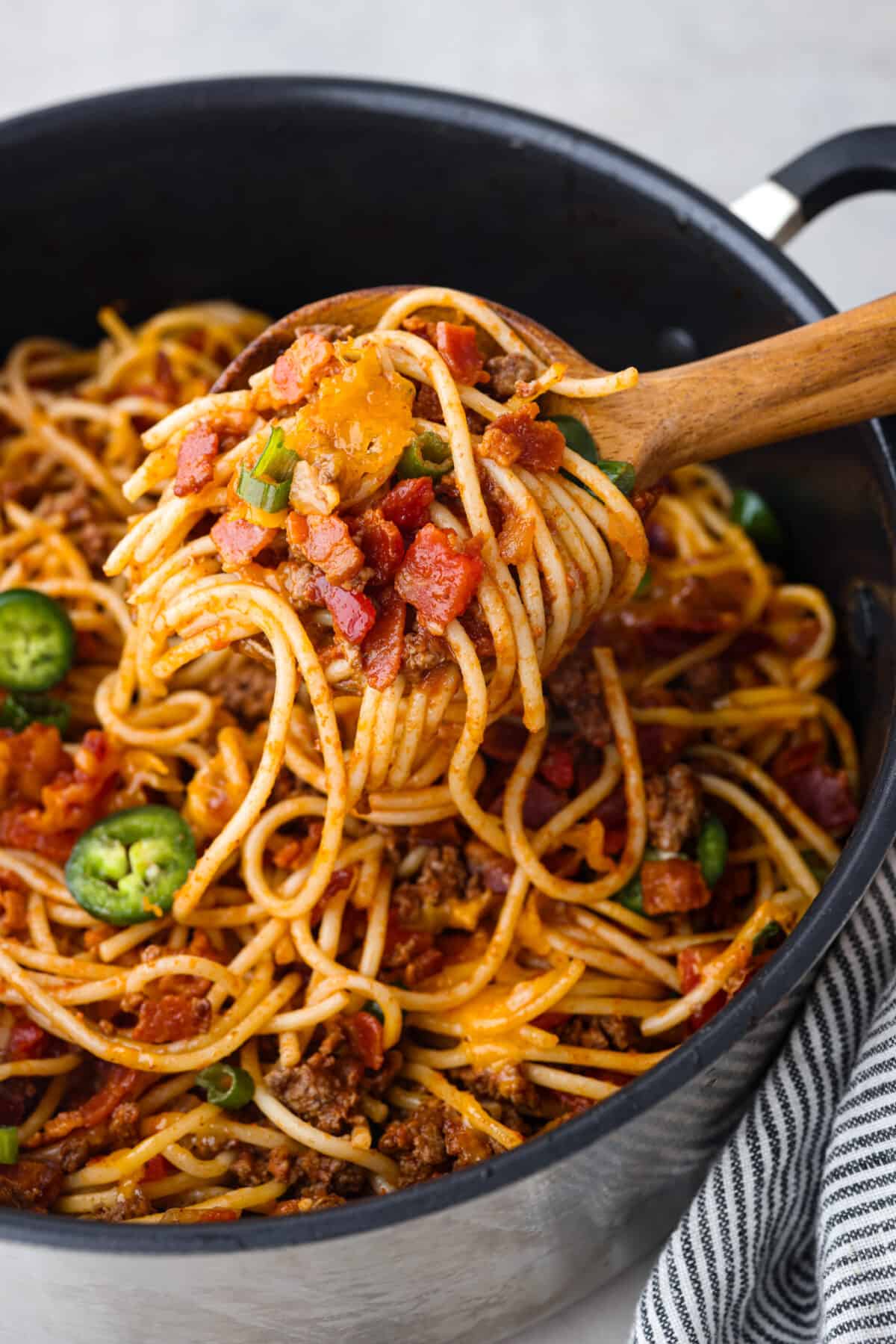 Upgrade your spaghetti with some cowboy fixings: sliced jalapenos, bacon, shredded cheddar cheese, and rotel tomatoes! It’s a hearty American version of a classic dish that everyone will love! A scoop of spaghetti on a wooden spoon.