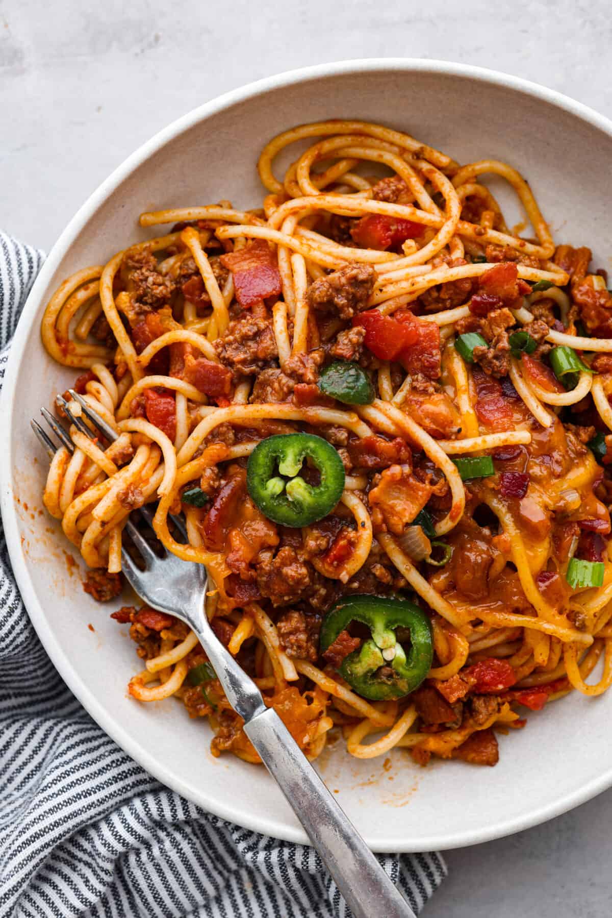 Upgrade your spaghetti with some cowboy fixings: sliced jalapenos, bacon, shredded cheddar cheese, and rotel tomatoes! It’s a hearty American version of a classic dish that everyone will love! A serving of Cowboy Spaghetti in a white bowl.