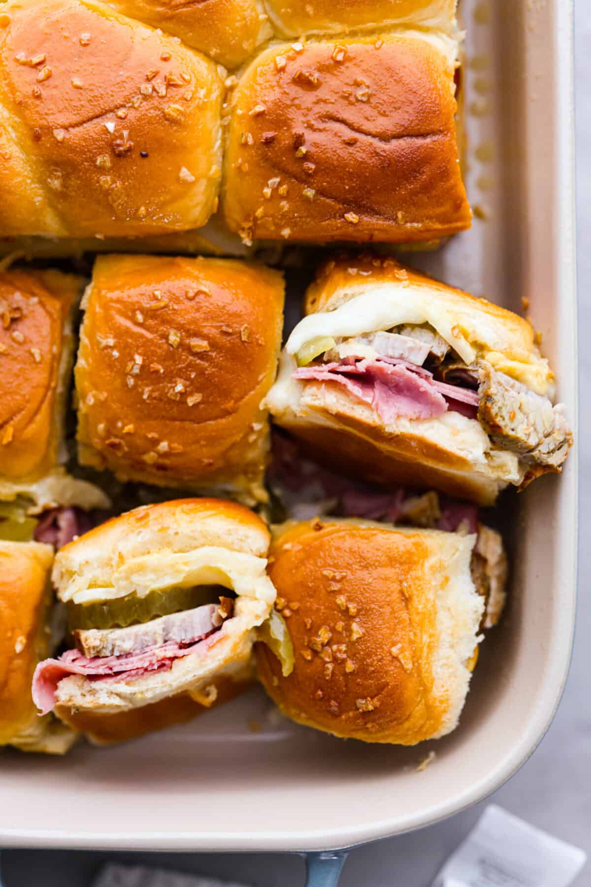 These Cuban Sliders are about to become your new go-to party dish! Think melty Swiss cheese, dill pickles, and all the meaty goodness of a Cuban sandwich, but bite-sized and perfect for sharing. Top-down view of the Cuban sliders in a white baking dish. Two are turned on their side so you can see the filling.