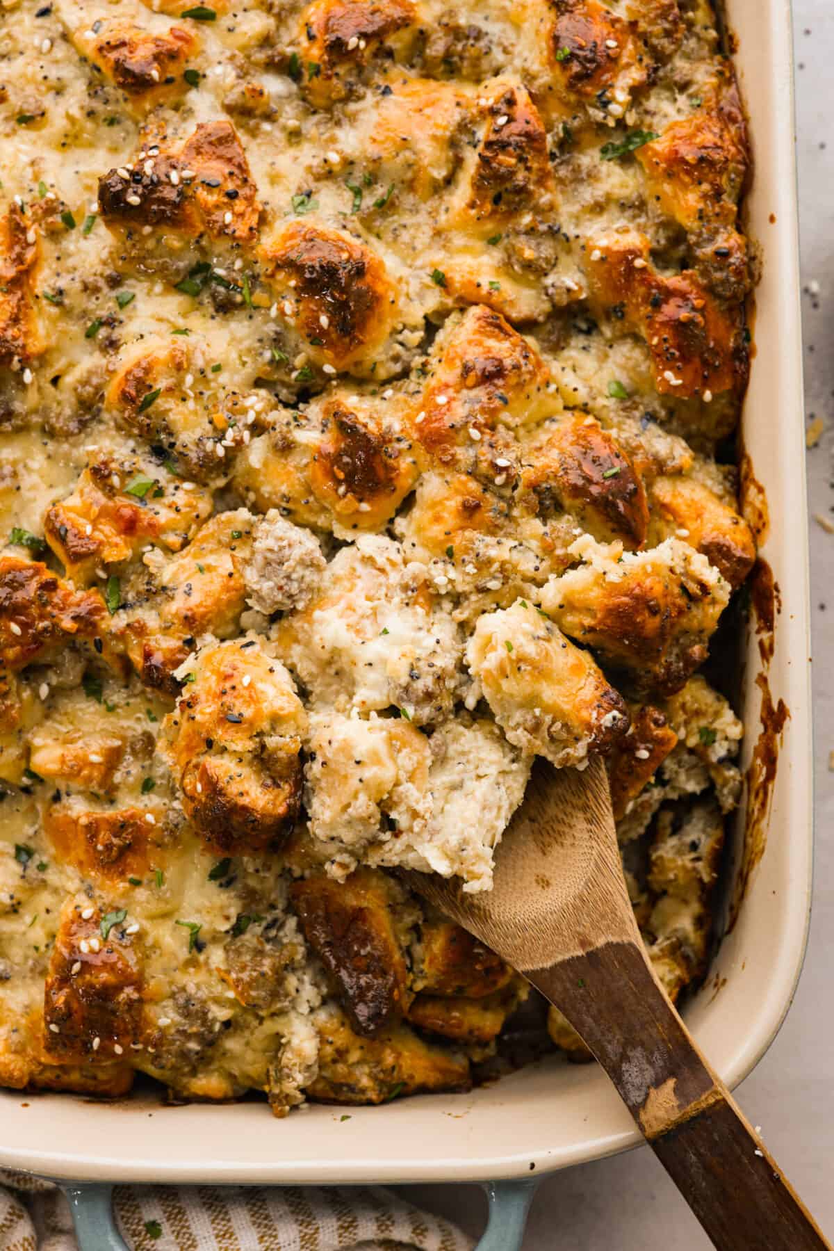 Wake up to the tasty flavors of this everything bagel casserole! It's made with everything bagels and seasoning, sausage, eggs, cream cheese, and gruyère cheese. The result is a creamy and savory breakfast casserole that everyone will devour! Close view of a wood spoon lifting up everything bagel casserole from the baking dish.