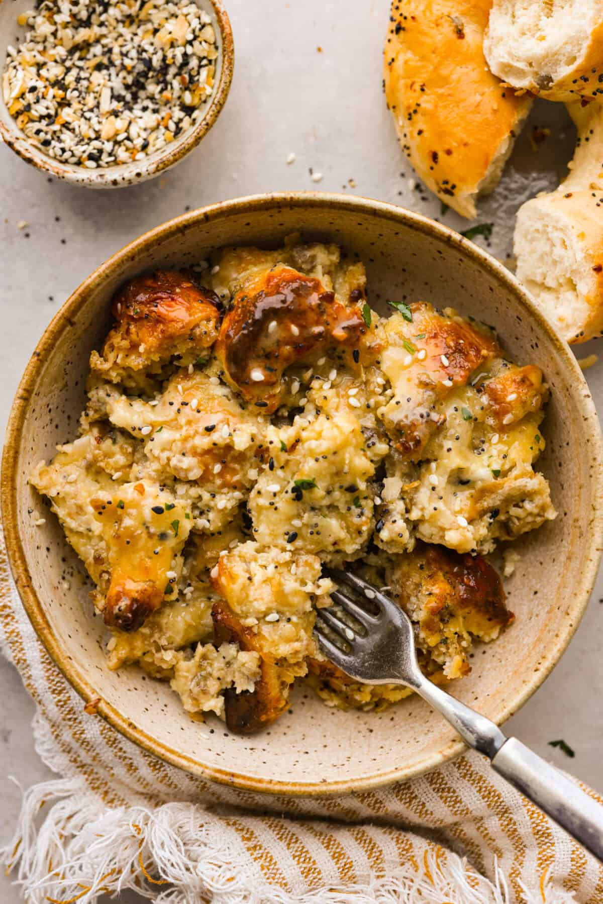 Wake up to the tasty flavors of this everything bagel casserole! It's made with everything bagels and seasoning, sausage, eggs, cream cheese, and gruyère cheese. The result is a creamy and savory breakfast casserole that everyone will devour! Top close view of everything bagel casserole in a brown bowl with a fork.