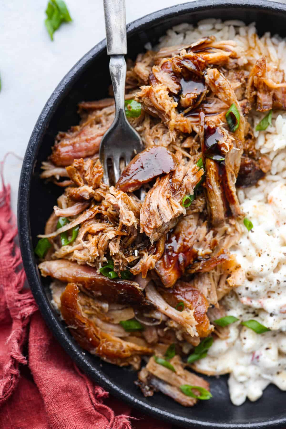 Tender slow cooker kalua pulled pork is salty, smokey melt-in-your-mouth perfection. Enjoy authentic Hawaiian luau pork flavor at home with this super simple 3 ingredient masterpiece. Top view of kalua pulled pork in a black bowl with rice and macaroni salad on the side. A fork is inside the bowl with teriyaki sauce and green onions garnished on top.