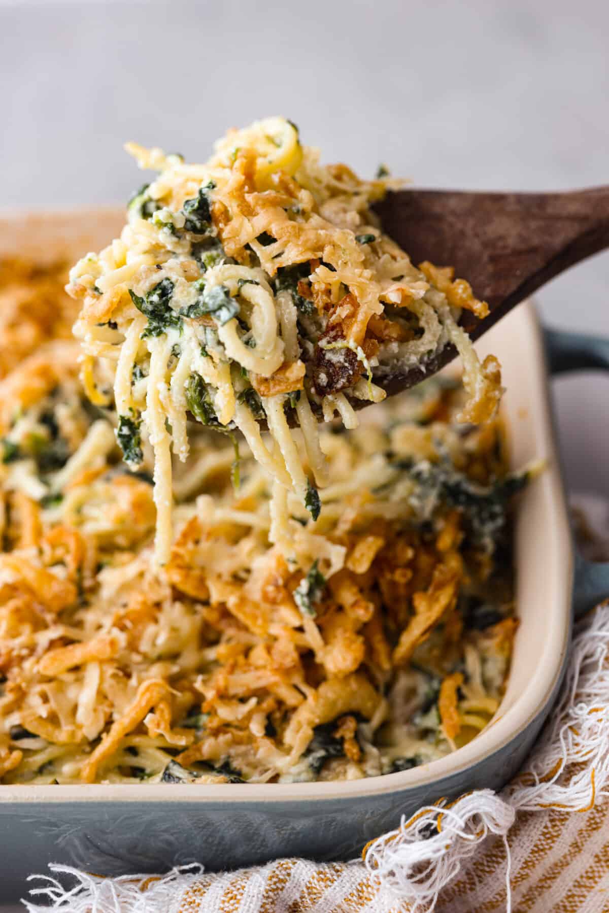 You will love the tasty flavors in this Monterey spaghetti! Tender spaghetti baked in a cheesy mixture and loaded with spinach and crispy fried onions. It's a surprisingly delicious combination of flavors and an easy weeknight dinner! Close view of a wood spoon lifting the spaghetti from the baking dish.