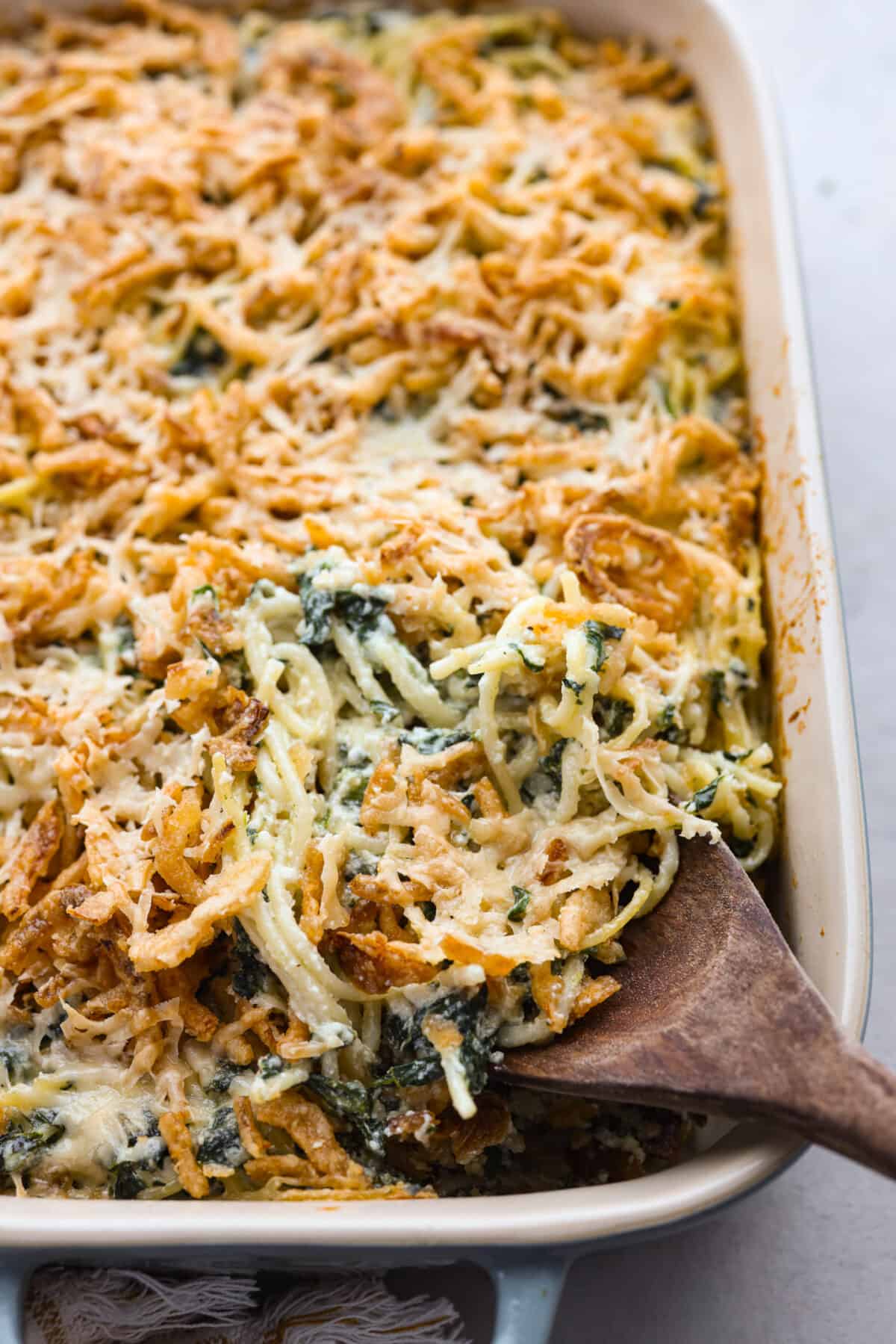 You will love the tasty flavors in this Monterey spaghetti! Tender spaghetti baked in a cheesy mixture and loaded with spinach and crispy fried onions. It's a surprisingly delicious combination of flavors and an easy weeknight dinner! Close view of Monterey spaghetti in a baking dish with a wooden serving spoon.