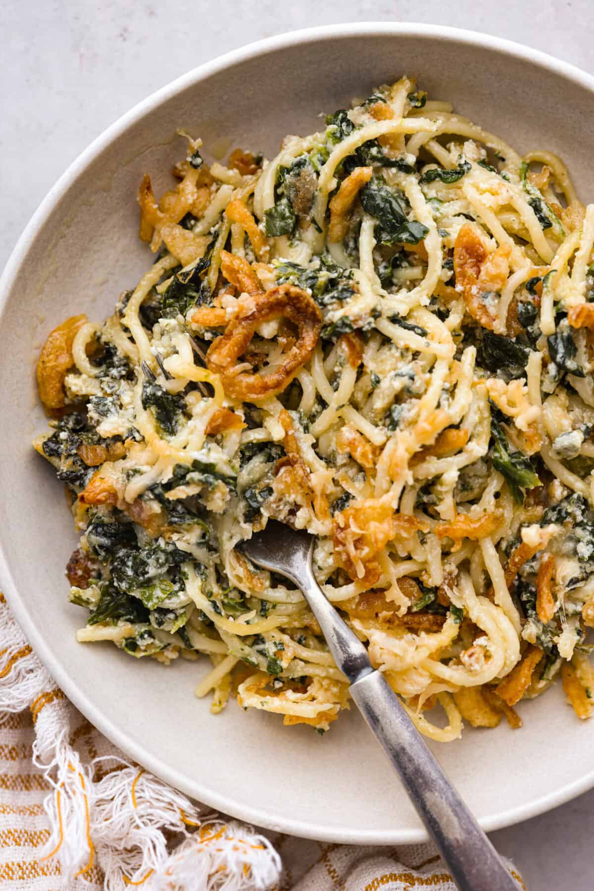 You will love the tasty flavors in this Monterey spaghetti! Tender spaghetti baked in a cheesy mixture and loaded with spinach and crispy fried onions. It's a surprisingly delicious combination of flavors and an easy weeknight dinner! Top view of Monterey spaghetti in a white bowl with a fork.