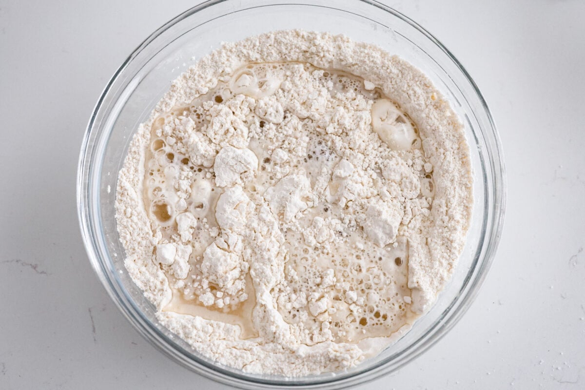 Make soft homemade bread without any kneading! This easy peasant bread recipe only uses 6 ingredients to make two perfect loaves of bread. Fluffy on the inside with a buttery golden crust on the outside. Second process photo of the wet ingredients added to the bowl of dry ingredients.