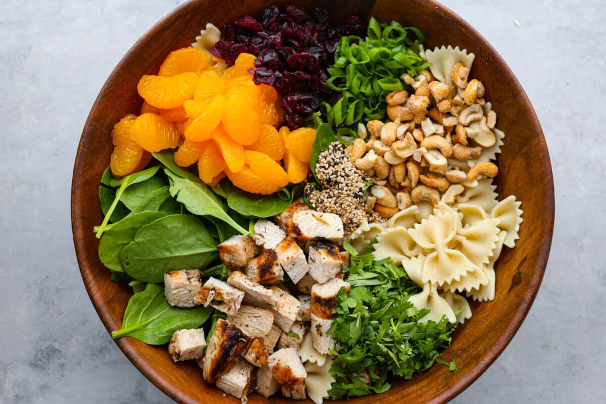 Teriyaki spinach pasta salad combines juicy mandarin oranges, savory teriyaki chicken, and crunchy cashews for a flavor explosion in every bite! The best Asian-inspired pasta salad to serve alongside your favorite meals. A wooden bowl filled with chicken, greens, mandarin oranges, bowtie pasta, and cashews.