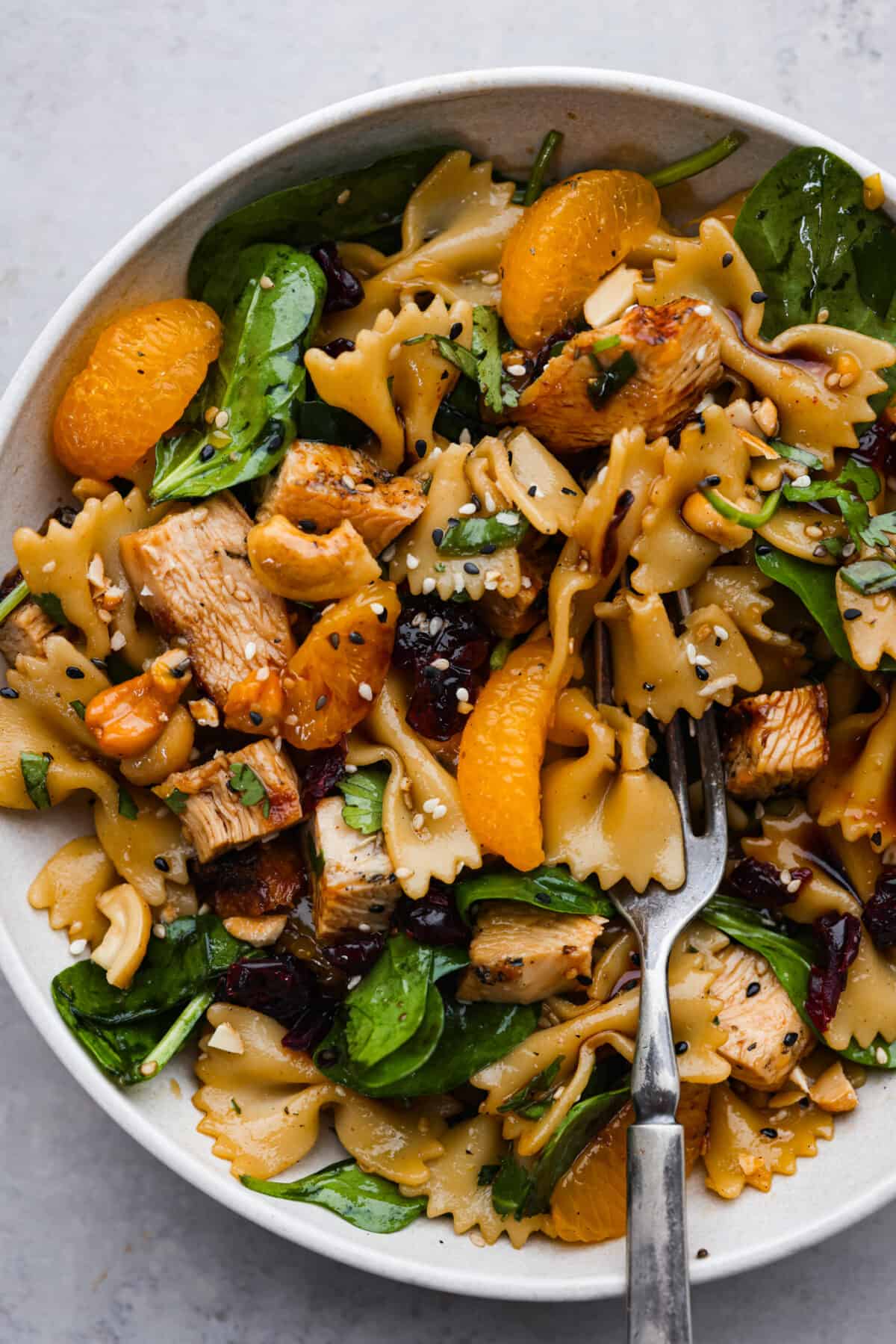Teriyaki spinach pasta salad combines juicy mandarin oranges, savory teriyaki chicken, and crunchy cashews for a flavor explosion in every bite! The best Asian-inspired pasta salad to serve alongside your favorite meals. A serving of teriyaki spinach pasta salad on a white plate.