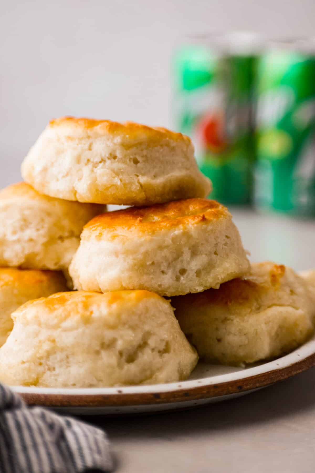 Tender and fluffyย 7-up biscuitsย rise to perfection and instantly become everyone's favorite biscuit! It only takes 4-ingredients to create this melt-in-your-mouth biscuit recipe! Close view of the 7 up biscuits stacked on a plate with 7 up in the back ground.