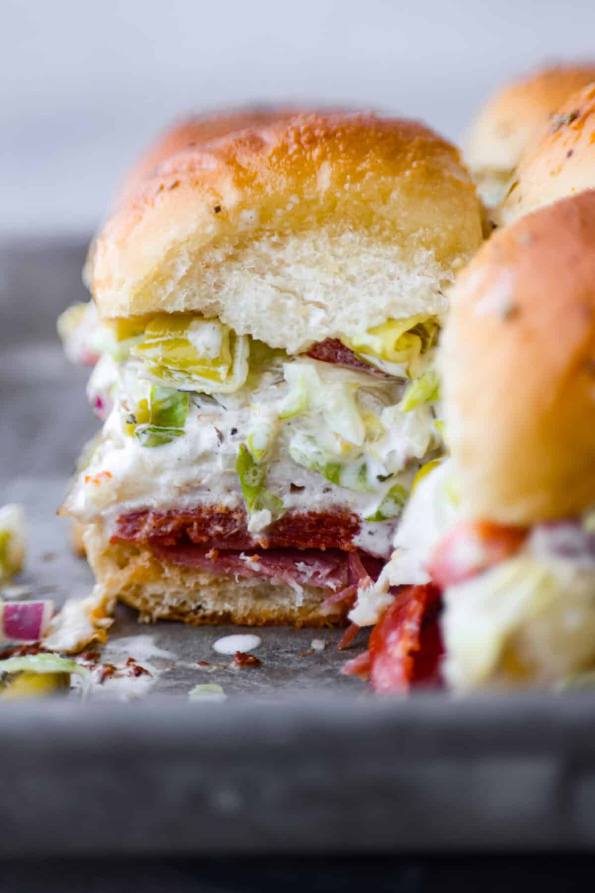 The classic grinder sandwich made into sliders to share! You've got Hawaiian rolls topped with layers of pepperoni, salami, turkey, provolone, and creamy grinder salad. And to top things off- each roll is brushed with a buttery parmesan glaze and baked to golden brown perfection. Closeup of a slider.