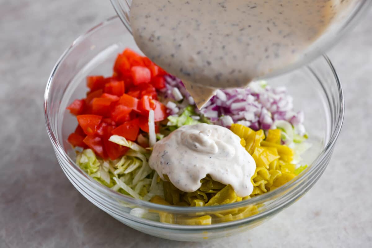 It’s like your favorite deli sub ditched the bread and got a glow-up. Think layers of crispy lettuce, your favorite meats, and a creamy dressing all in this yummy Grinder Salad form! Someone pouring dressing over salad ingredients.