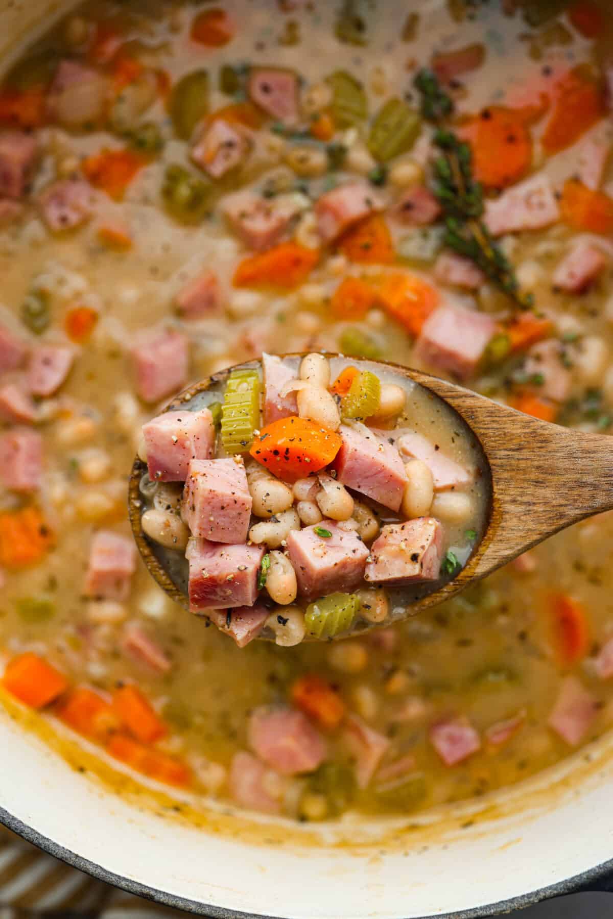 Comforting ham and bean soup is a classic and hearty soup. It's loaded with creamy white beans, smoky ham, and tender veggies. It's the perfect soup for using up your leftover ham and super easy to make on those busy weeknights. Close view of a wooden spoon lifting up a spoonful of ham and bean soup.