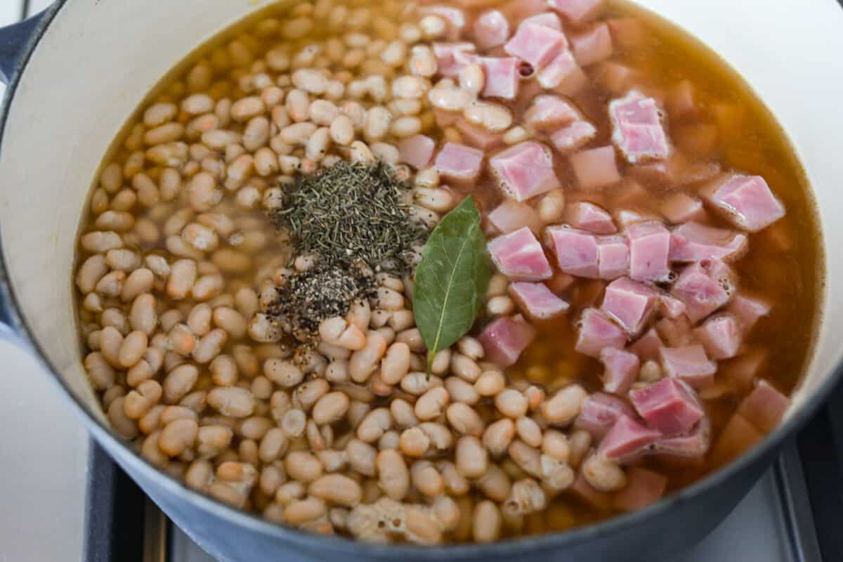 Comforting ham and bean soup is a classic and hearty soup. It's loaded with creamy white beans, smoky ham, and tender veggies. It's the perfect soup for using up your leftover ham and super easy to make on those busy weeknights. Fourth process photo of the bay leaf, pepper, and thyme added to the pot.