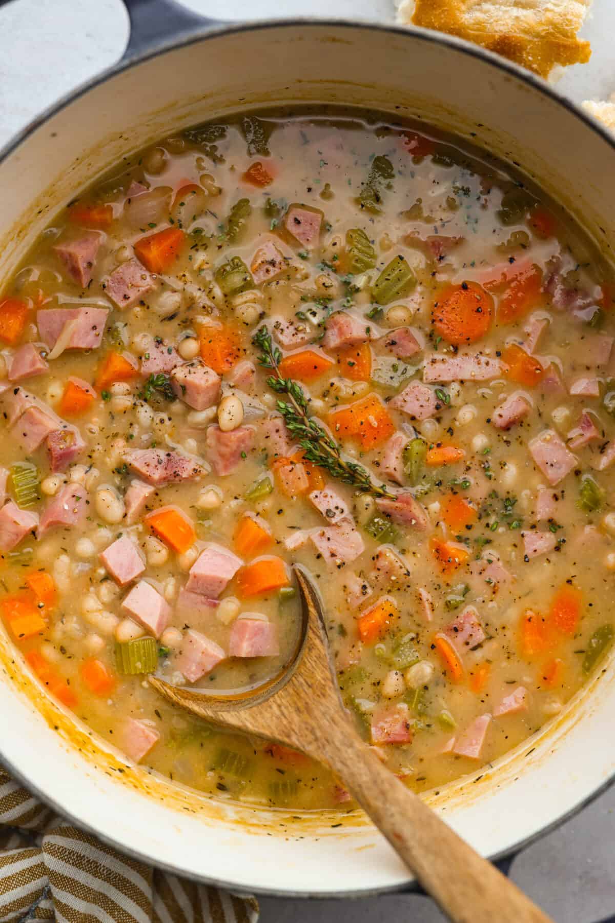 Comforting ham and bean soup is a classic and hearty soup. It's loaded with creamy white beans, smoky ham, and tender veggies. It's the perfect soup for using up your leftover ham and super easy to make on those busy weeknights. Top view of ham and bean soup in a large stock pot with a wooden spoon and garnished with thyme.