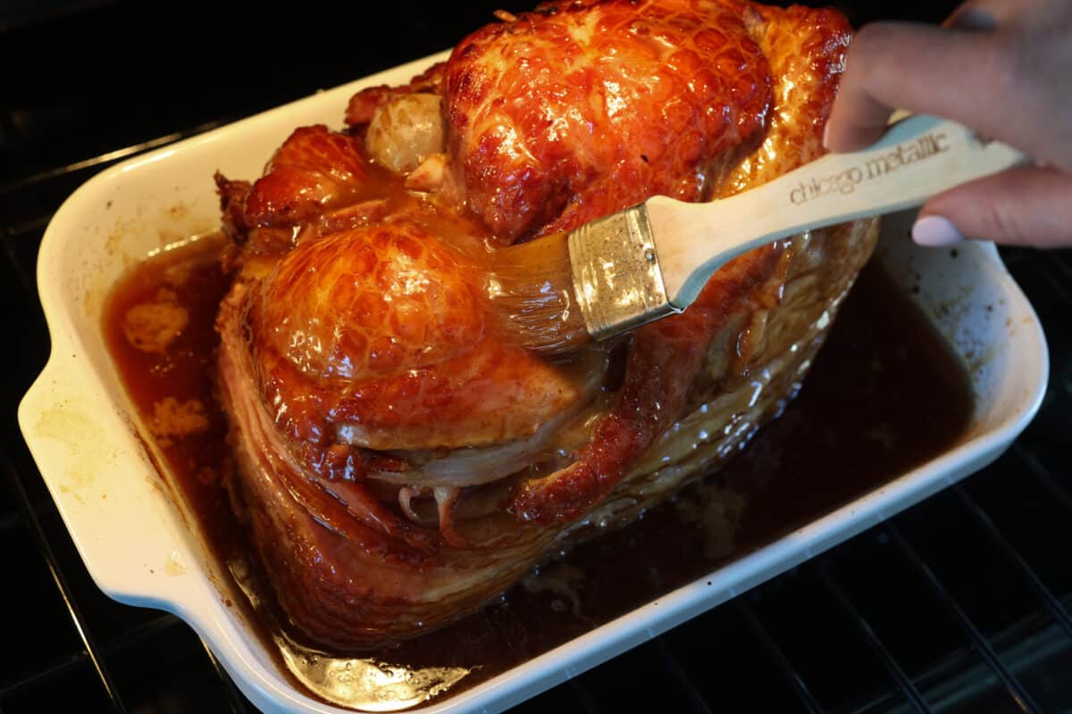 This honey-glazed ham is coated with delicious brown sugar and sweet honey glaze! It’s tender and juicy and goes with everything which makes it ideal for any holiday meal! A honey baked ham with more glaze being added to it.