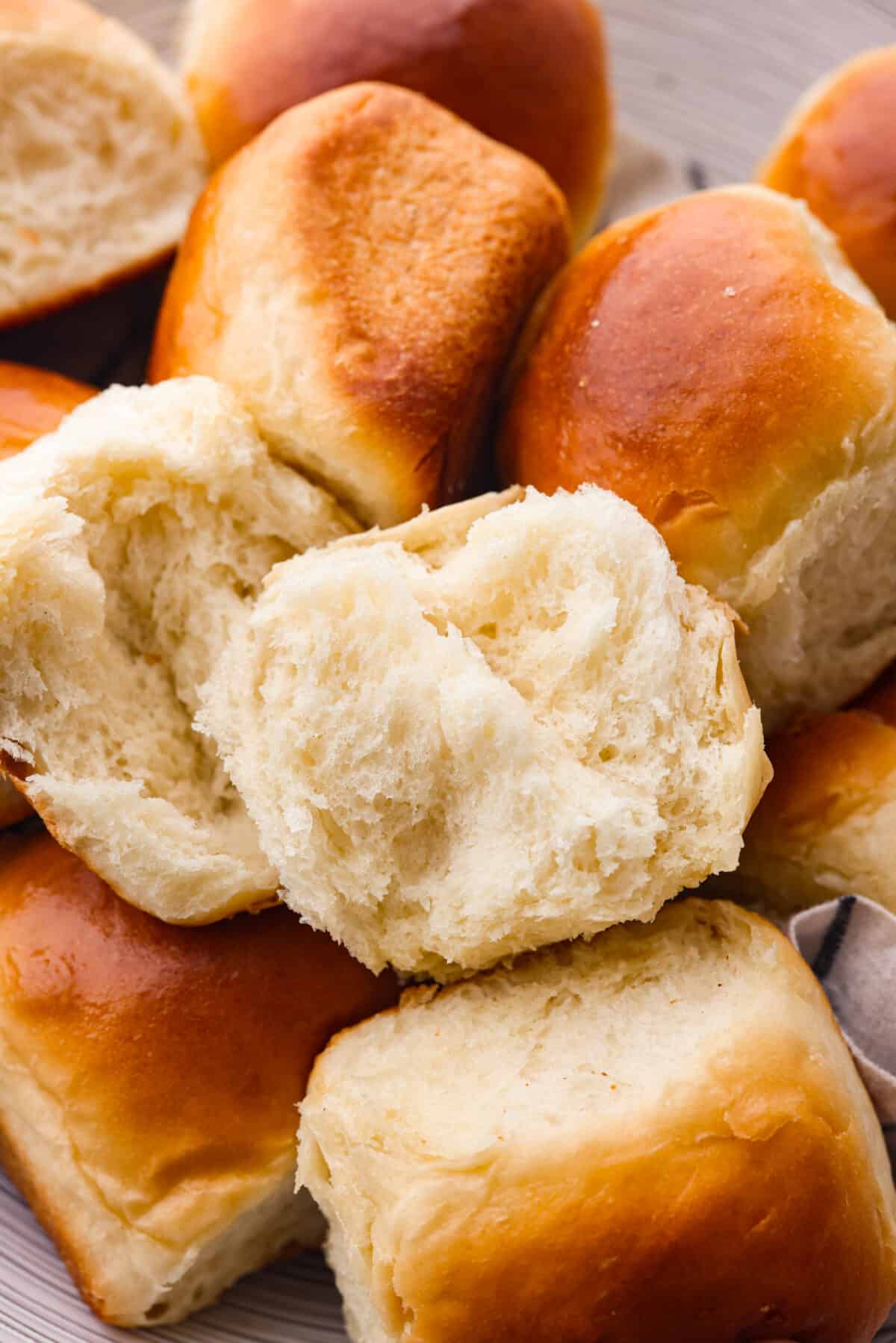 Perfectly soft buttery rolls will be your classic go-to roll recipe! They are tender fluffy, and baked with a buttery golden top. These rolls are sure to be a family favorite! Close view of a perfect soft and buttery roll split open on top of a pile of rolls.