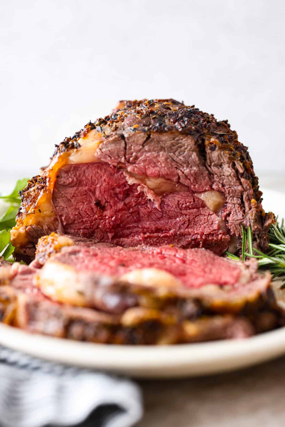 Tender and juicy ribeye roast is an incredibly succulent cut of beef. Seasoned with a garlic herb butter rub and cooked to the perfect melt-in-your-mouth temperature. An impressive and delicious roast made easy! Close view of ribeye roast on a platter with herbs.