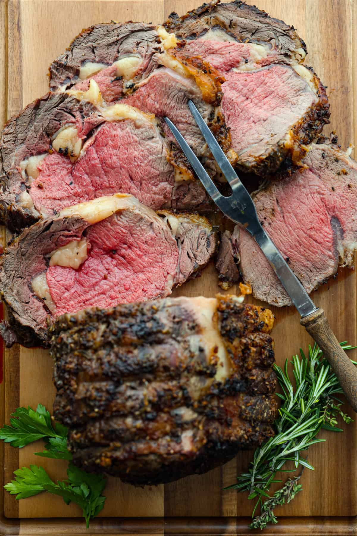 Tender and juicy ribeye roast is an incredibly succulent cut of beef. Seasoned with a garlic herb butter rub and cooked to the perfect melt-in-your-mouth temperature. An impressive and delicious roast made easy! Top view of sliced ribeye roast on a wood cutting board with a large serving fork.