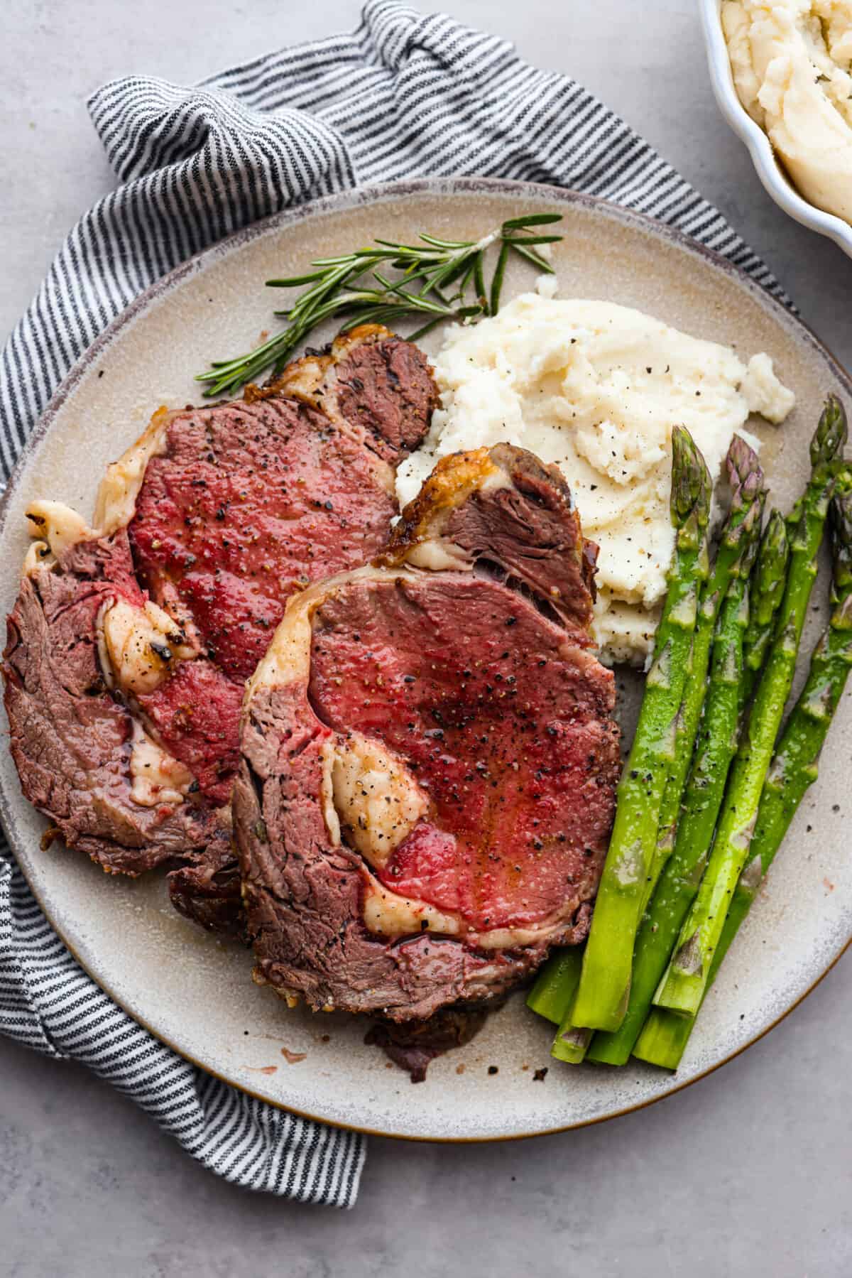 Tender and juicy ribeye roast is an incredibly succulent cut of beef. Seasoned with a garlic herb butter rub and cooked to the perfect melt-in-your-mouth temperature. An impressive and delicious roast made easy! Top view of sliced ribeye roast on a large plate with mashed potatoes and asparagus on the side.