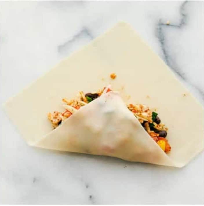 These are the BEST Southwest chicken egg rolls! It's aย colorfulย combination of corn, black beans, fresh veggies, shredded cheese, and chicken. This tasty mixture is flavored in savory southwestย seasonings then fried in a crispy egg roll wrapper. These won't last long at your next party! Second photo of the bottom corner of the wrapper folding over the mixture.