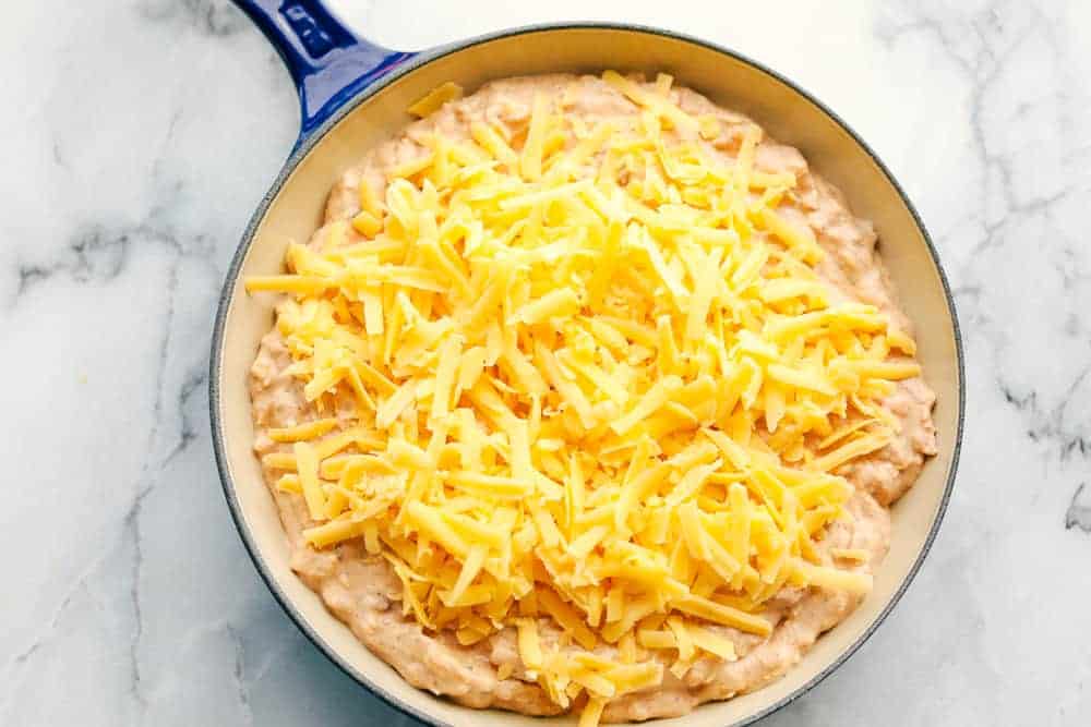 Bring out the chips and start dipping because this is the most addicting bean dip you will ever have! It's creamy, cheesy, and loaded with tasty Mexican-inspired flavors. Garnish with toppings, and take this to your next gathering! Process shot of bean dip in a blue skillet with cheese on top before baking.
