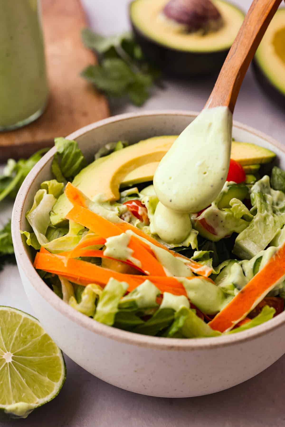 This dressing is packed with flavor and so creamy! It's delicious on salads, burrito bowls, tacos, and even burgers and wraps. I love how quick and easy it is to make with simple ingredients, and it has the perfect balance of tangy, nutty and subtly sweet flavors. Side shot of a bowl of salad with veggies, avocado slices with a wooden spoon drizzling dressing over the top.