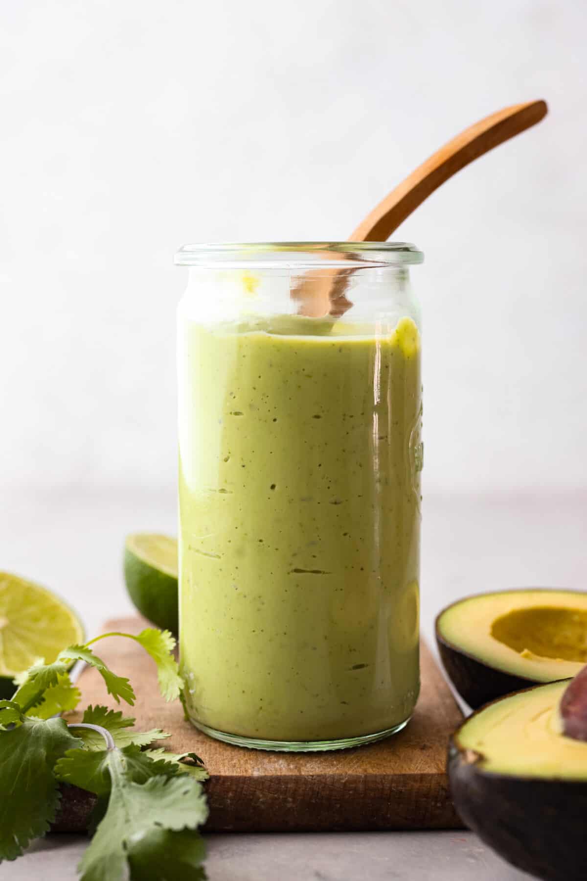 This dressing is packed with flavor and so creamy! It's delicious on salads, burrito bowls, tacos, and even burgers and wraps. I love how quick and easy it is to make with simple ingredients, and it has the perfect balance of tangy, nutty and subtly sweet flavors. Side view of a glass jar filled with avocado dressing and a wooden spoon sticking out. There are avocados, limes and cilantro on the table next to the jar.