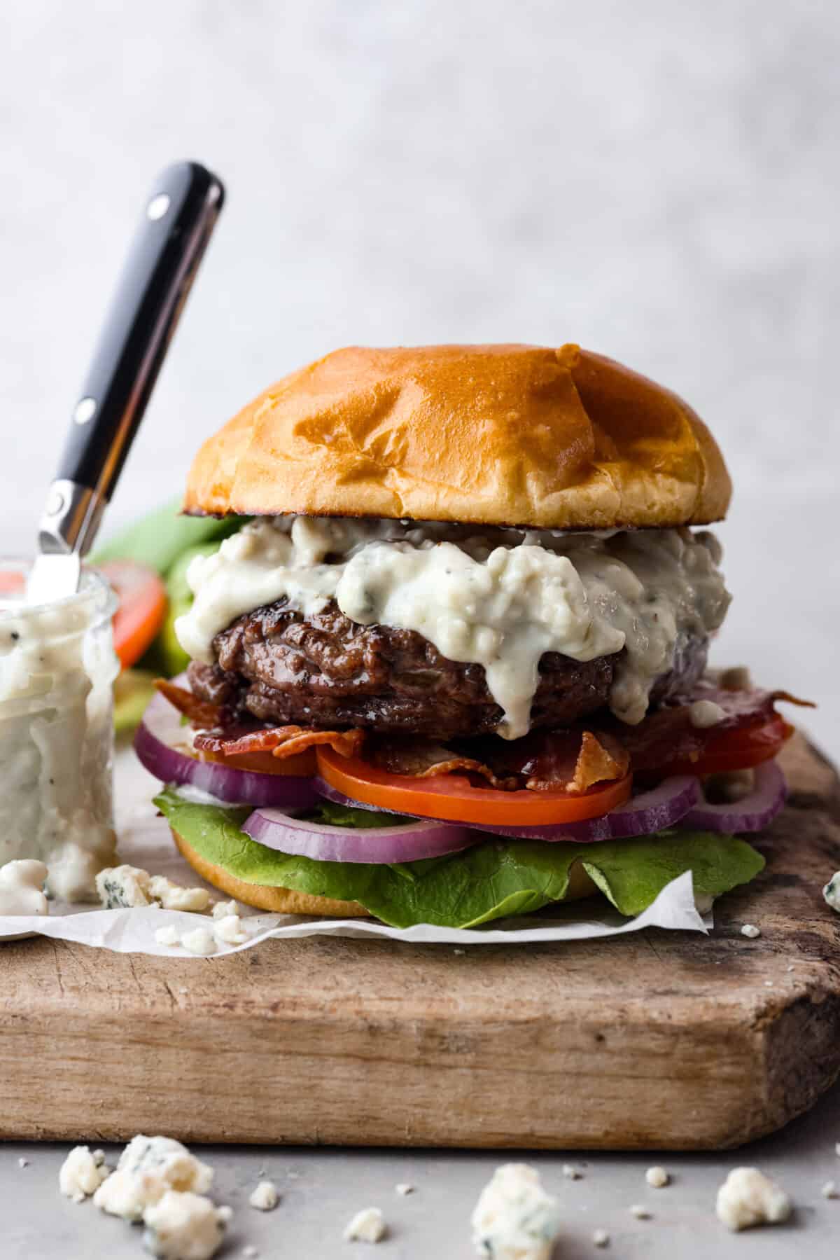 Get ready to be blown away by this Blue Cheese Burger! It's thick, juicy, and filled with flavor. If you love blue cheese dressing, this recipe is for you! That unique tangy flavor is the perfect addition to the juiciest patty ever. Side view of a burger with blue cheese dressing, bacon, lettuce tomato and onion on a crispy bun. All atop a wood cutting board with blue cheese crumbles.
