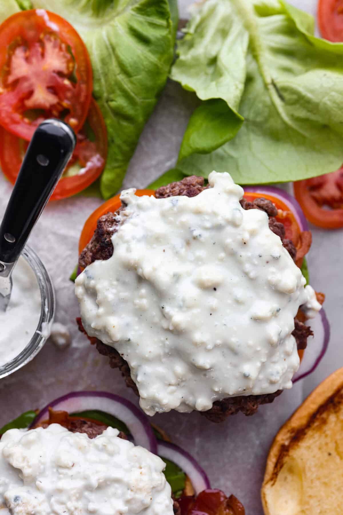 Get ready to be blown away by this Blue Cheese Burger! It's thick, juicy, and filled with flavor. If you love blue cheese dressing, this recipe is for you! That unique tangy flavor is the perfect addition to the juiciest patty ever. Overhead shot of burgers without top bun loaded with blue cheese dressing.