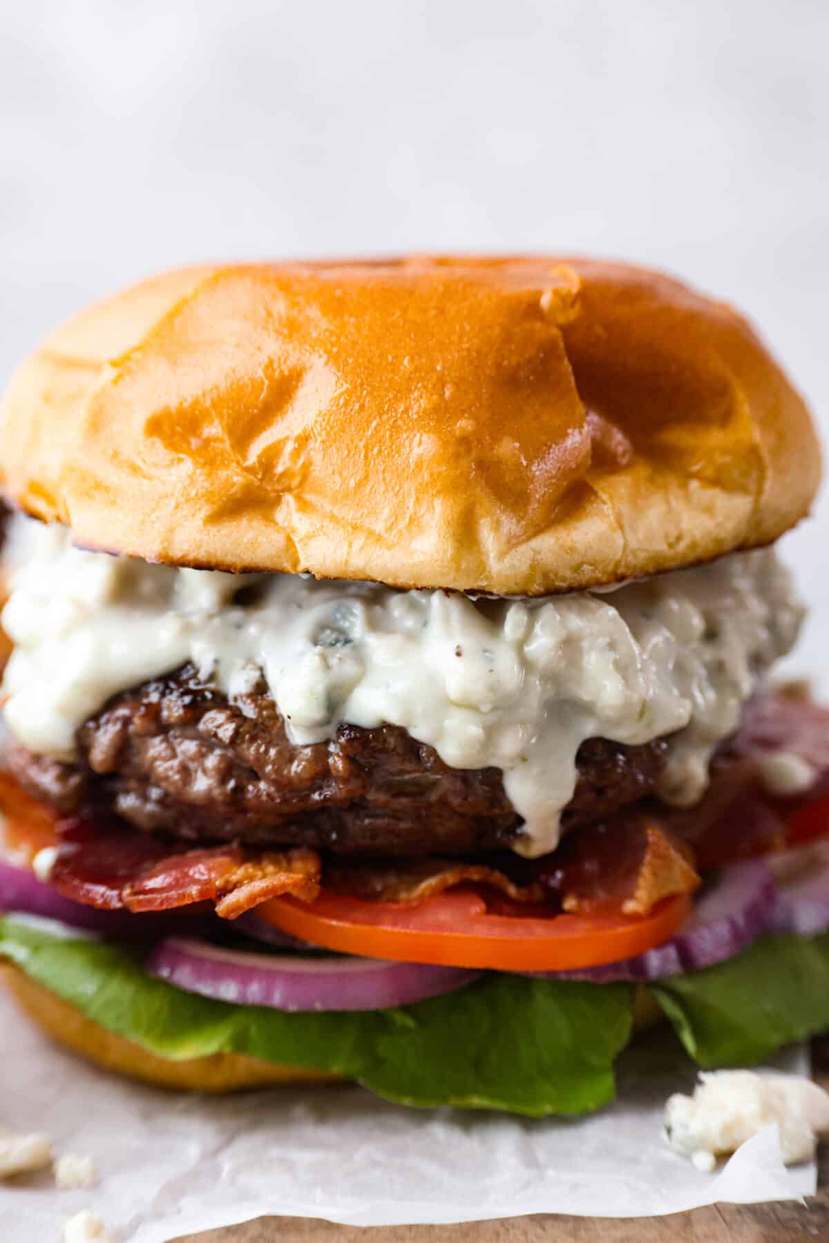 Get ready to be blown away by this Blue Cheese Burger! It's thick, juicy, and filled with flavor. If you love blue cheese dressing, this recipe is for you! That unique tangy flavor is the perfect addition to the juiciest patty ever. Close up side view of burger dripping with blue cheese dressing, toasted bun, lettuce, tomato, bacon and red onion.