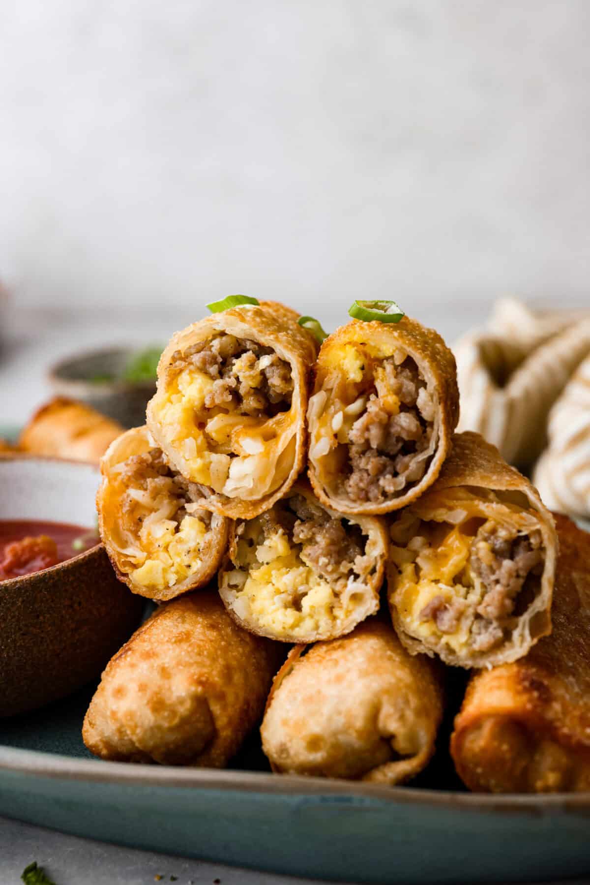 Breakfast Egg Rolls are a fun and delicious way to change up breakfast! The inside is filled with savory sausage, creamy eggs, hash browns, and plenty of cheese. Fry them up for a gooey, tender, savory center and a delicately crunchy exterior. This is a fun and unique breakfast that everyone will love! Side shot of stacked breakfast egg rolls, cut through middle so you can see the inside.
