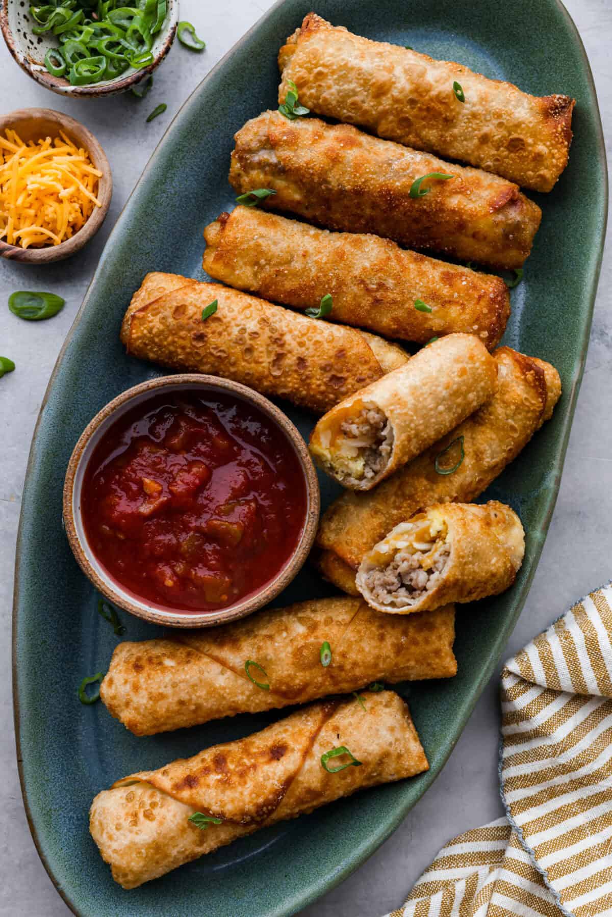 Breakfast Egg Rolls are a fun and delicious way to change up breakfast! The inside is filled with savory sausage, creamy eggs, hash browns, and plenty of cheese. Fry them up for a gooey, tender, savory center and a delicately crunchy exterior. This is a fun and unique breakfast that everyone will love! Overhead shot of a plate of cooked egg rolls with salsa in a bowl next to egg rolls.