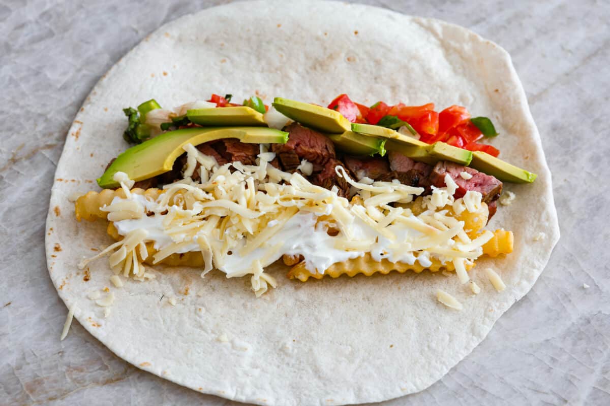 This delicious California Burrito is filled with well-seasoned grilled steak, pepper jack cheese, French fries, pico de Gallo, sour cream and avocado, all wrapped up in a warm flour tortilla. A flavorful lunch or dinner option that will be an absolute hit! A tortilla being filled to make a California burrito.