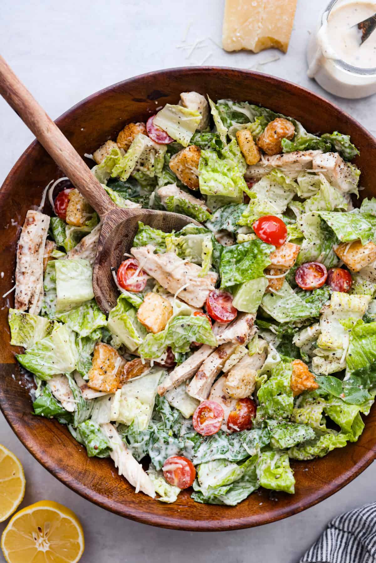Fresh and delicious Grilled Chicken Caesar Salad is a crowd-pleasing hearty salad with tender grilled chicken, crisp romaine lettuce, cherry tomatoes, shredded parmesan cheese, and crunchy croutons! Top view of a chicken caesar salad in a wooden bowl with a wooden spoon in it.