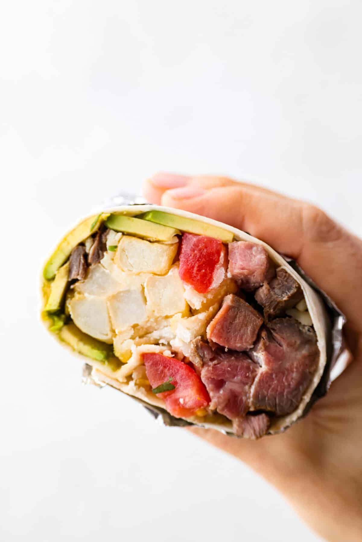 This delicious California Burrito is filled with well-seasoned grilled steak, pepper jack cheese, French fries, pico de Gallo, sour cream and avocado, all wrapped up in a warm flour tortilla. A flavorful lunch or dinner option that will be an absolute hit! A half holding a California burrito that is sliced open so that you can see the inside.