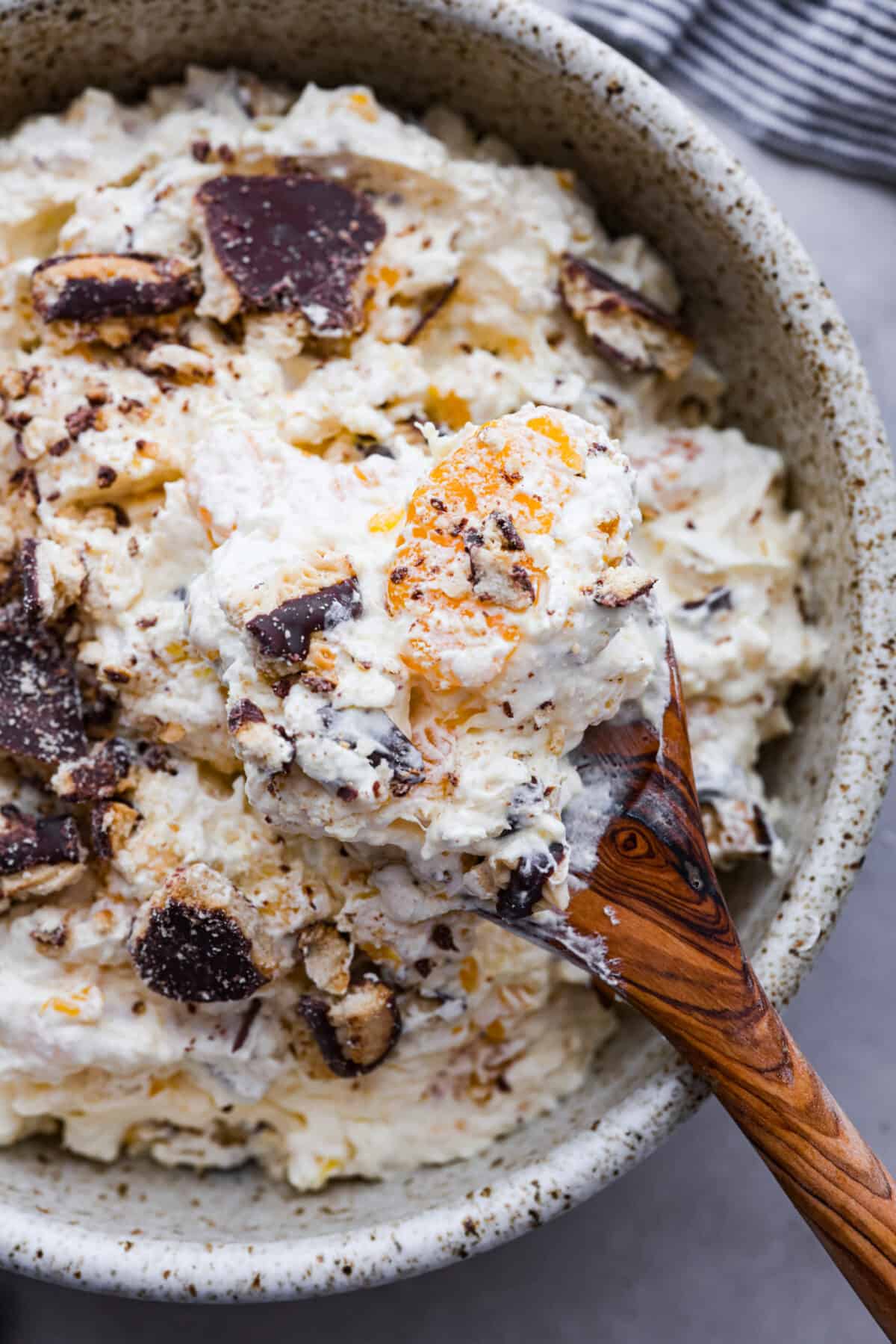 Serve this fun and delicious cookie salad as a dessert or side dish! Everyone will love the sweet fluffy pudding base mixed with mandarin oranges, crushed pineapple, and fudge stripes cookies. Close view of a wood serving spoon lifting up a spoonful of cookie salad.