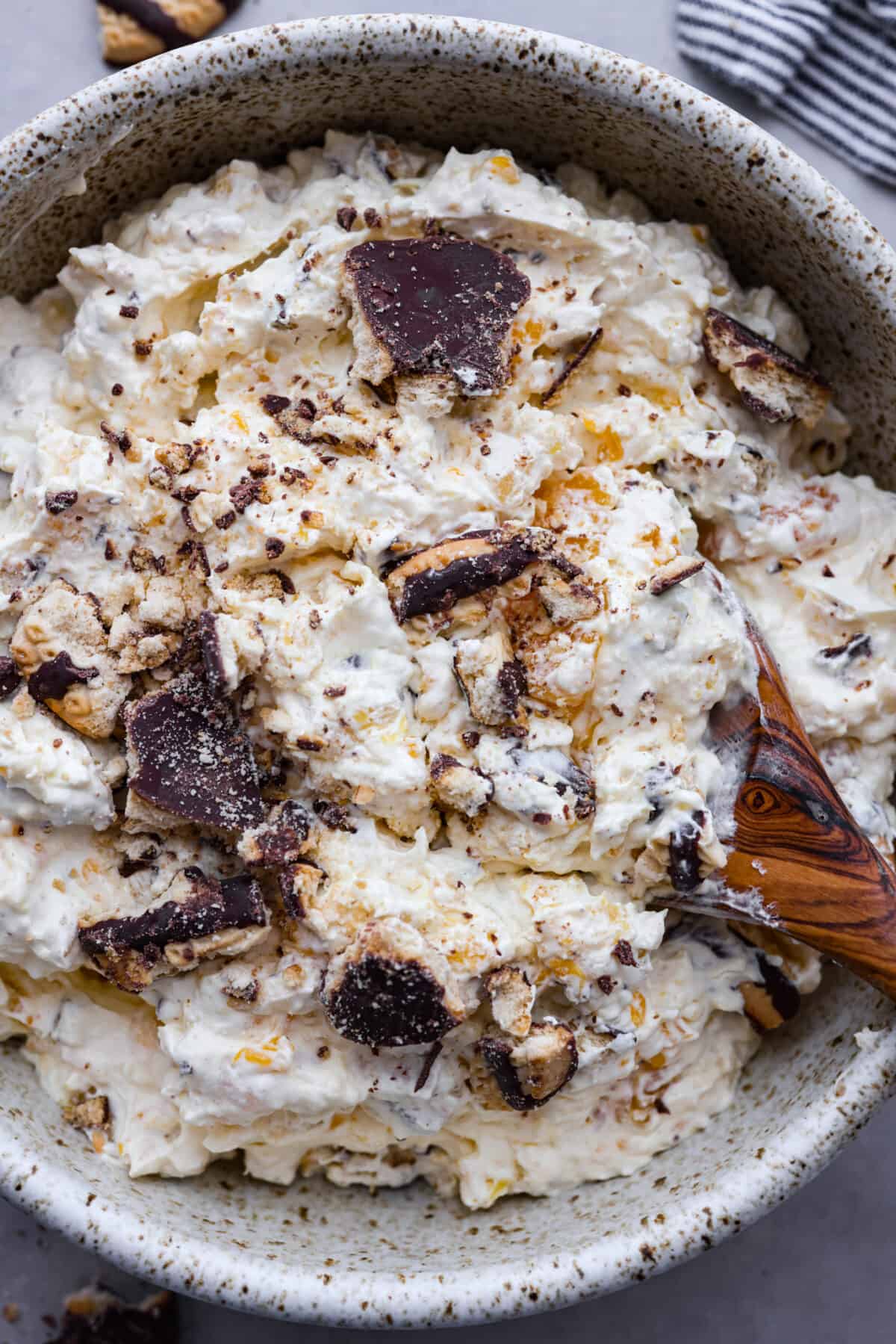 Serve this fun and delicious cookie salad as a dessert or side dish! Everyone will love the sweet fluffy pudding base mixed with mandarin oranges, crushed pineapple, and fudge stripes cookies. Top close view of cookie salad in a bowl with a wood serving spoon.