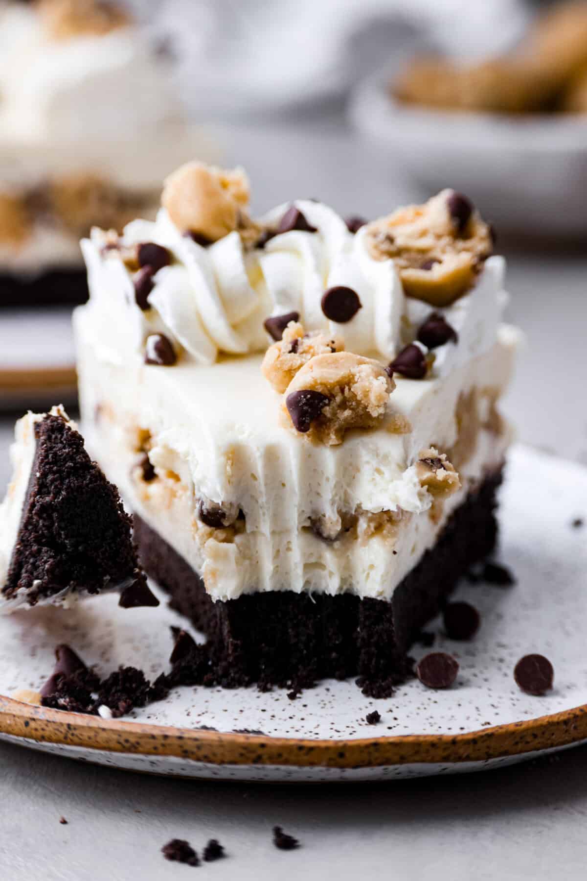 I am so excited about this recipe, it combines two of my all-time favorite desserts, cookie dough and cheesecake! With the rich Oreo crust, creamy cheesecake filling, and edible cookie dough bites, this no-bake cookie dough cheesecake will have your taste buds begging for more! Close-up image of the front of a slice of the no-bake cookie dough cheesecake with a bite taken out of it which is sitting on a fork next to the slice.