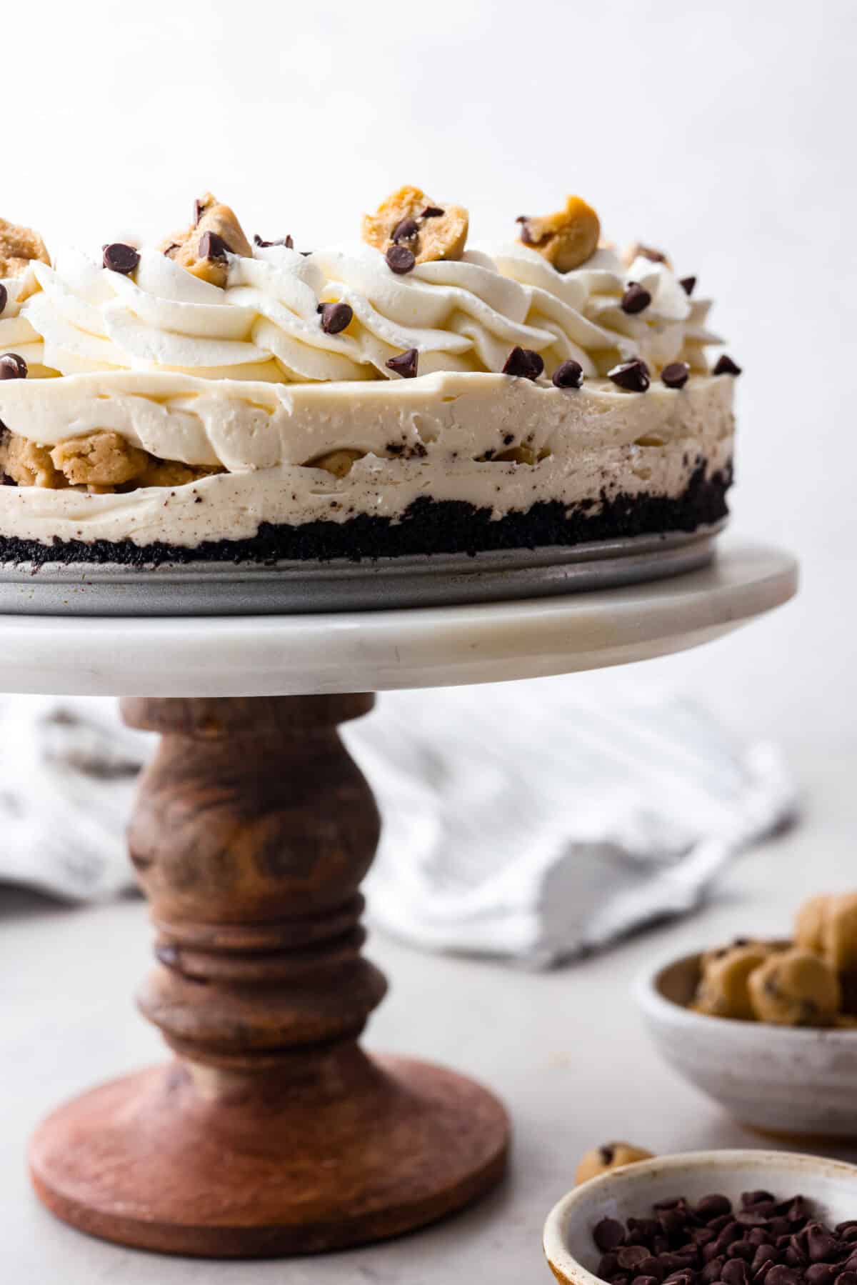 I am so excited about this recipe, it combines two of my all-time favorite desserts, cookie dough and cheesecake! With the rich Oreo crust, creamy cheesecake filling, and edible cookie dough bites, this no-bake cookie dough cheesecake will have your taste buds begging for more! Side shot of completed no-bake cookie dough cheesecake on a cake stand. you can see all the layers and toppings.