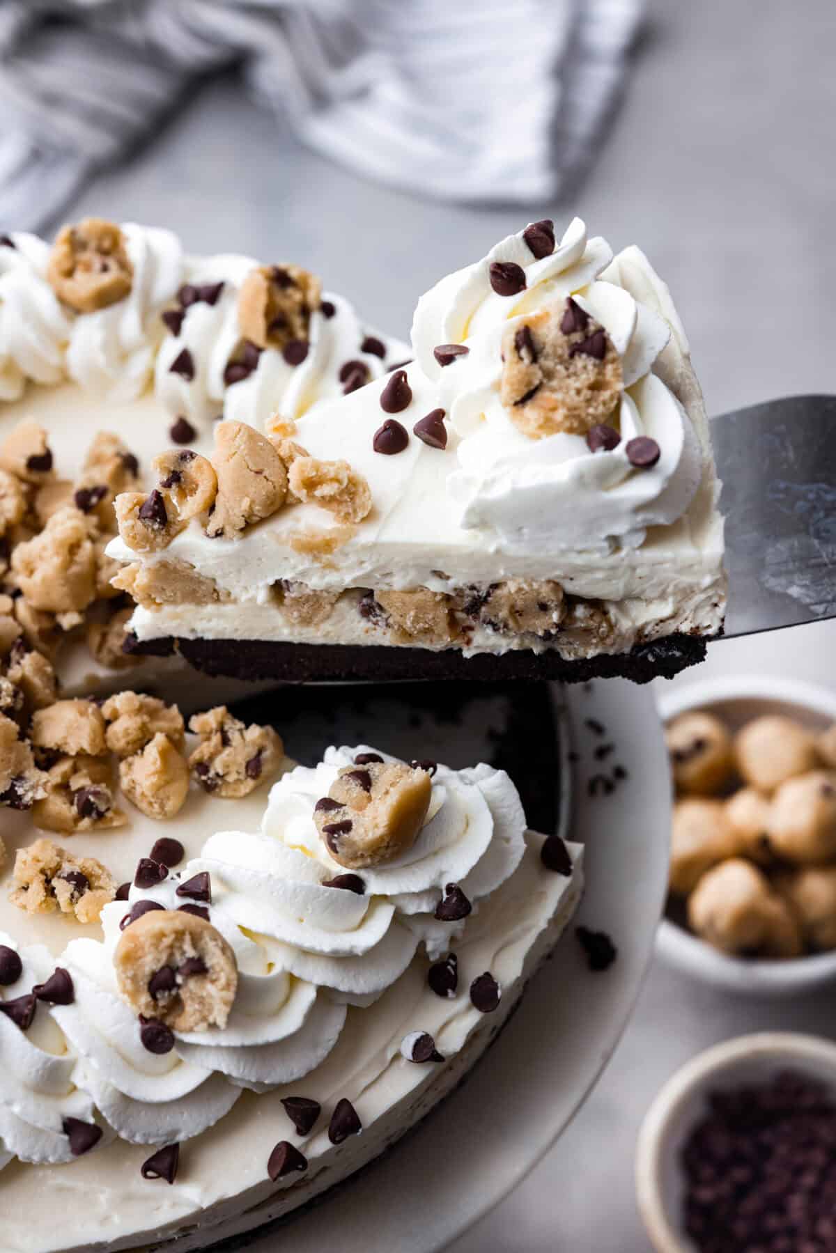 With the rich Oreo crust, creamy cheesecake filling, and edible cookie dough bites, this no-bake cookie dough cheesecake will have your taste buds begging for more! Slice of no-bake cookie dough cheesecake being taken out from the rest of the cheesecake. There is an Oreo crust, layers of creamy cheesecake filling and bits of cookie dough. Topped with more pieces of cookie dough mini chocolate chips and whipped cream.