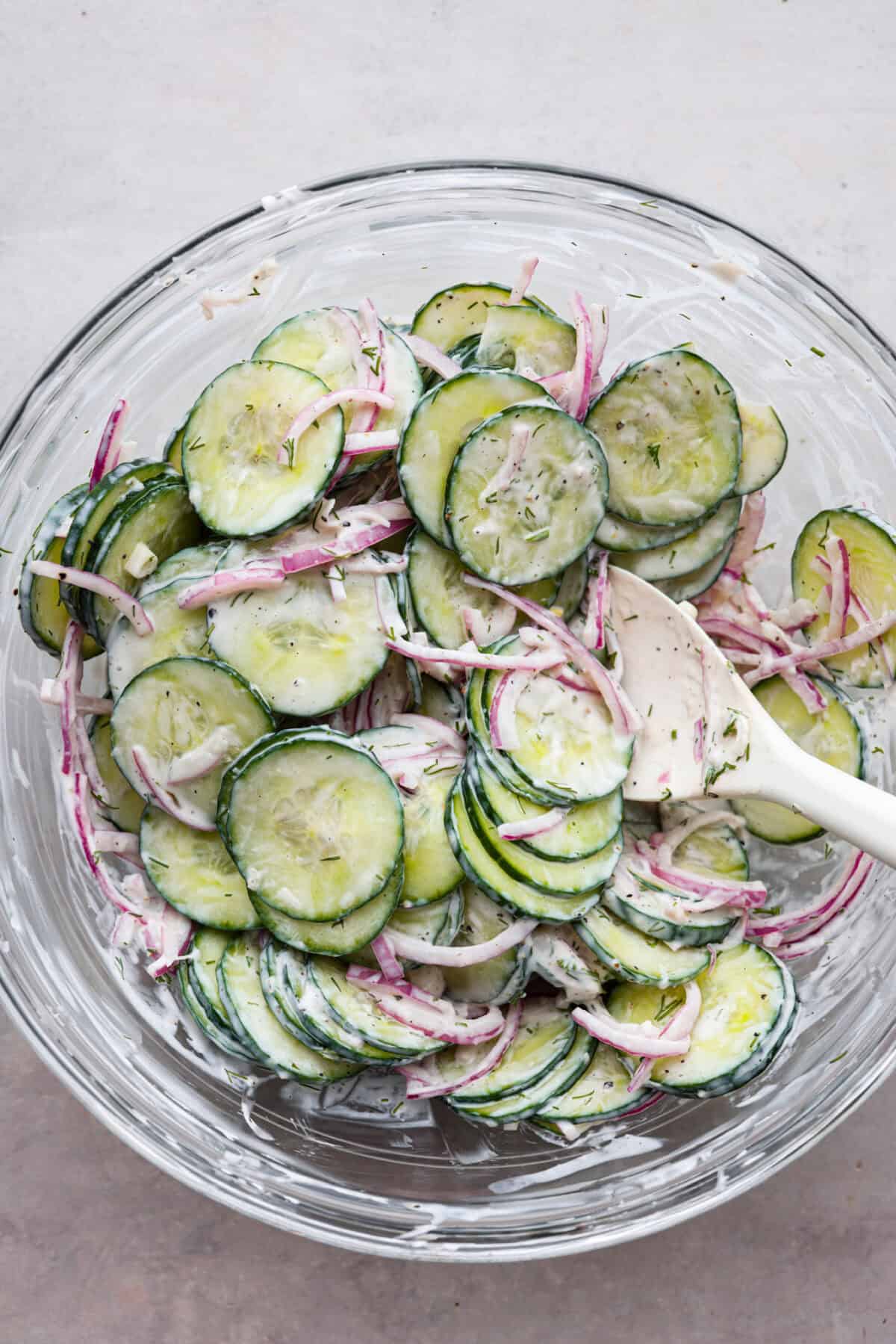 Creamy Cucumber Salad is a delicious and refreshing side dish. All it takes is a handful of fresh simple ingredients and 15 minutes. This is the perfect addition to your next summer barbecue! All of the ingredients for creamy cucumber salad being stirred in a glass bowl.